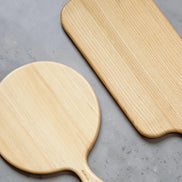 Ecohub NZ Entertainers Ash Round Paddle Board (35x25cm) - natural ash wood - lying next to a rectangular board on grey concrete